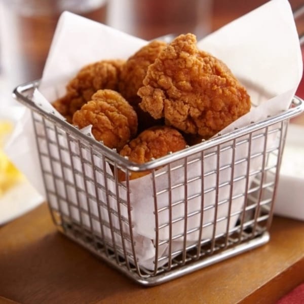 Crispy fried chicken pieces in a metal basket lined with white paper, on a wooden surface.